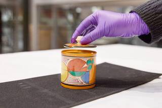 A gloved hand takes the lid off a ceramic jar